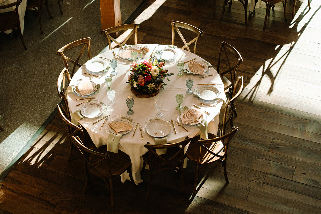 Round table set for eight with white tablecloth, plates, glasses, cutlery, and a floral centerpiece, surrounded by wooden chairs on a wooden floor—perfect for a Spring Woodland Wedding at Stone Valley Meadows.