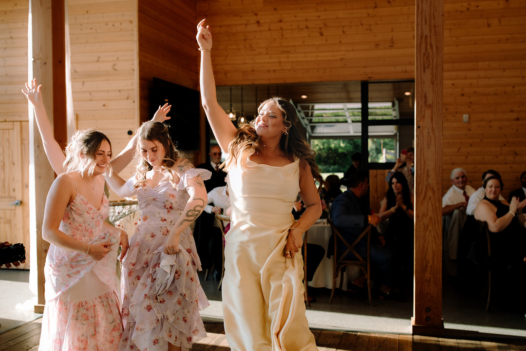 Three women in dresses dance together indoors at a sunlit Stone Valley Meadows event, while seated guests watch in the background, capturing the joy of a Spring Woodland Wedding celebration.