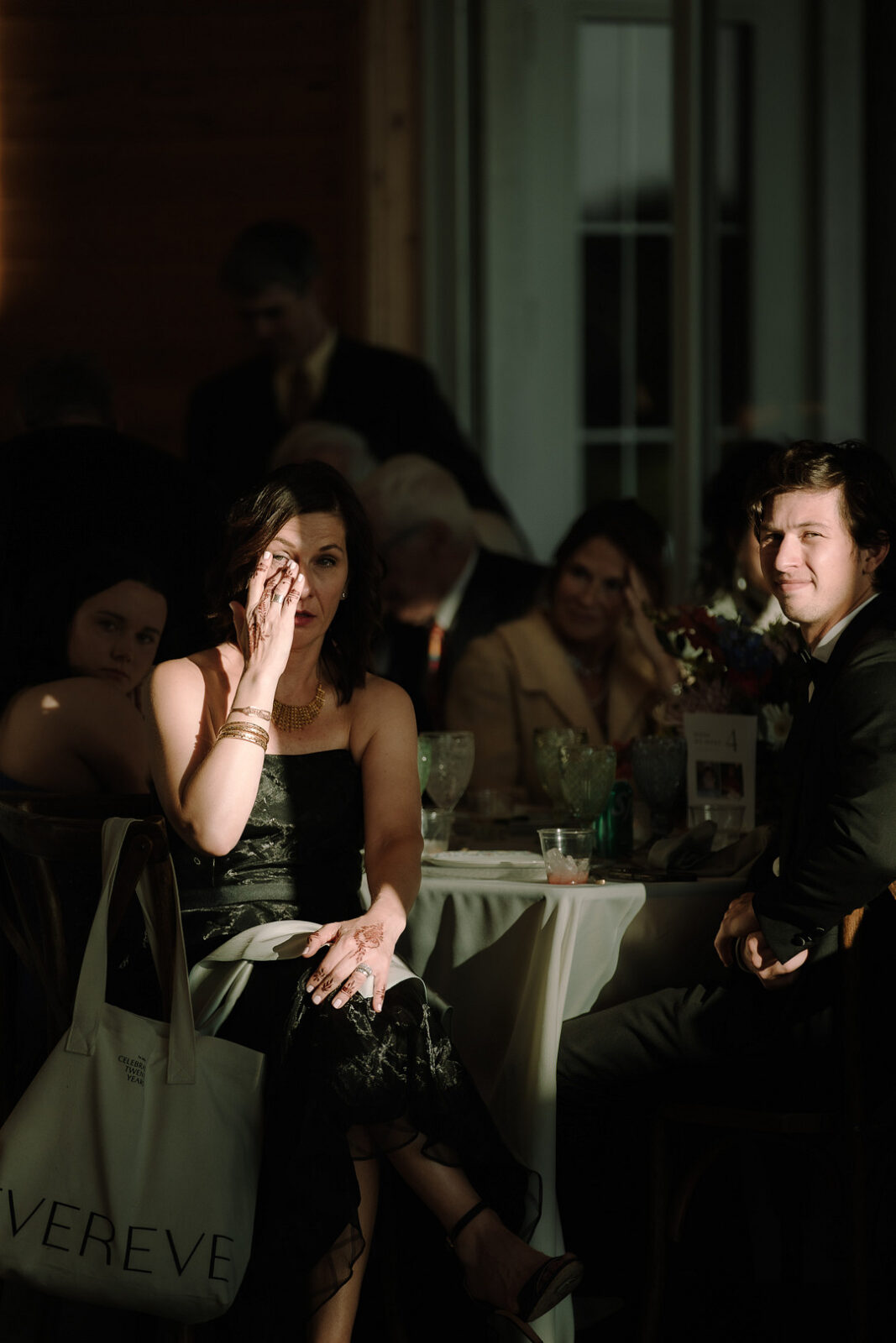 A woman in a black dress wipes her eye while sitting at a table during a Spring Woodland Wedding at Stone Valley Meadows. Other attendees are seated nearby, with sunlight partially illuminating the heartfelt scene.