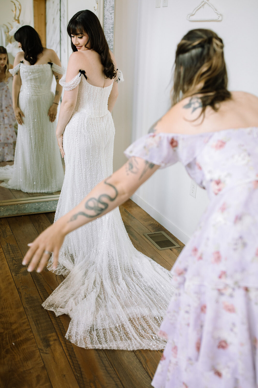 A bride in a white gown looks at her reflection in a mirror at Stone Valley Meadows, while another woman in a floral dress adjusts the train of the dress, capturing the magic of a Spring Woodland Wedding.