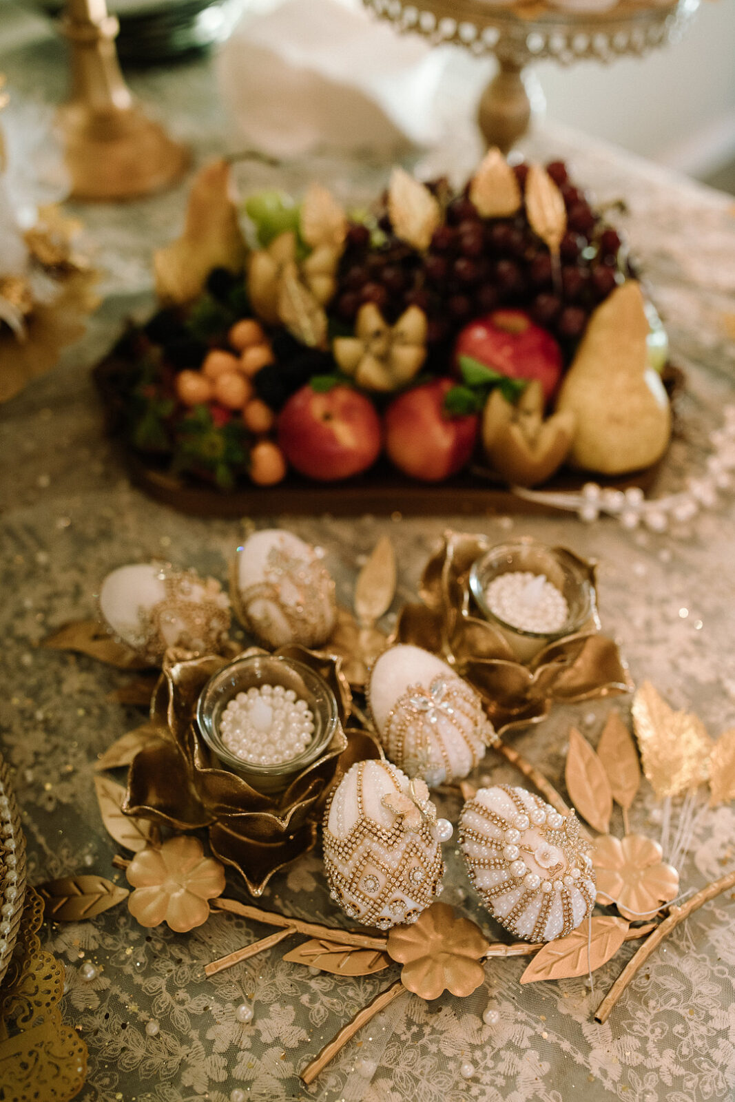 A decorative table setting for a Spring Woodland Wedding at Stone Valley Meadows, featuring ornate beaded eggs, gold floral accents, and a platter of assorted fresh fruits on a lace tablecloth.