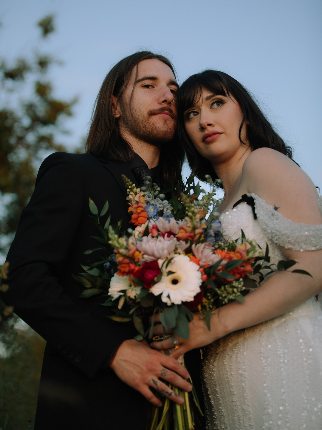 A bride in a white dress and a groom in a black suit stand close together outdoors at Stone Valley Meadows, holding a colorful bouquet—perfect for a Spring Woodland Wedding.