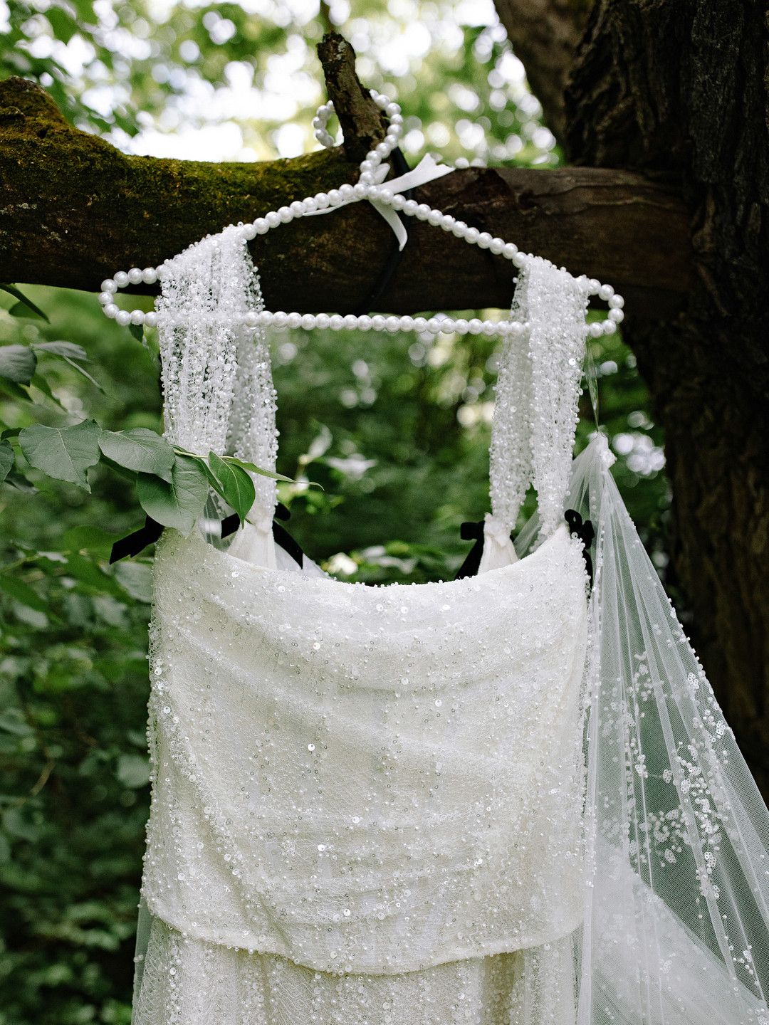 A white beaded wedding dress and veil hang on a pearl-covered hanger from a tree branch, surrounded by green foliage—a dreamy scene perfect for a Spring Woodland Wedding at Stone Valley Meadows.