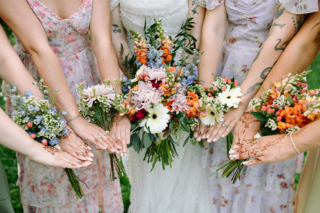 A group of women in floral dresses hold bouquets and display intricate henna designs on their hands, celebrating a Spring Woodland Wedding at Stone Valley Meadows.