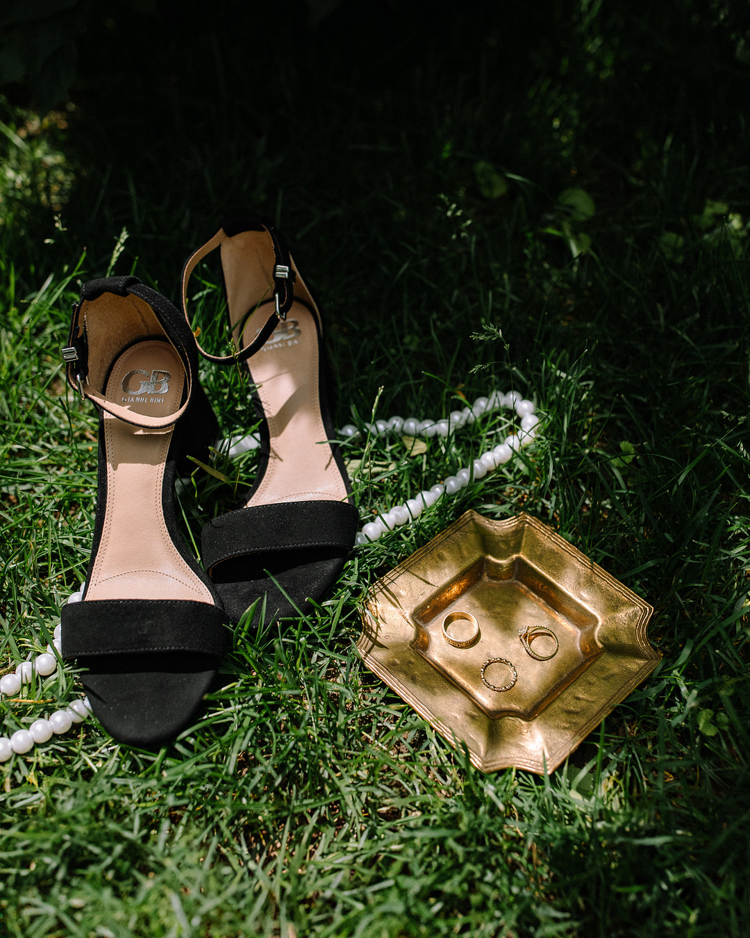 A pair of black high-heeled sandals, a pearl necklace, and a gold tray with rings and a bracelet are artfully arranged on green grass—perfect details for a Spring Woodland Wedding at Stone Valley Meadows.