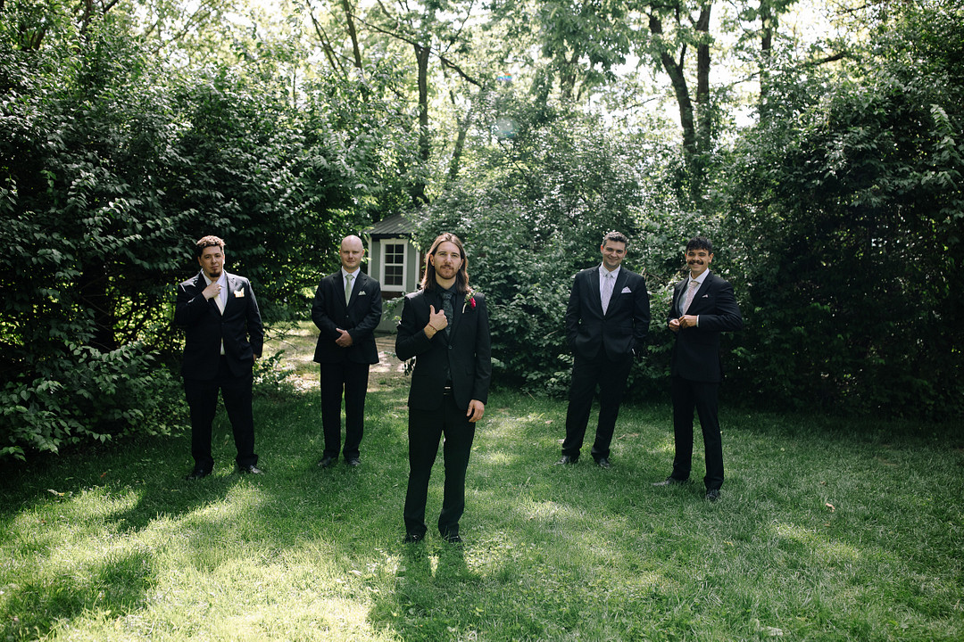 Five men in suits stand on grass in a sunlit, leafy yard at Stone Valley Meadows; one man stands in front, making a gesture with his hand, while the others pose behind him at this Spring Woodland Wedding.