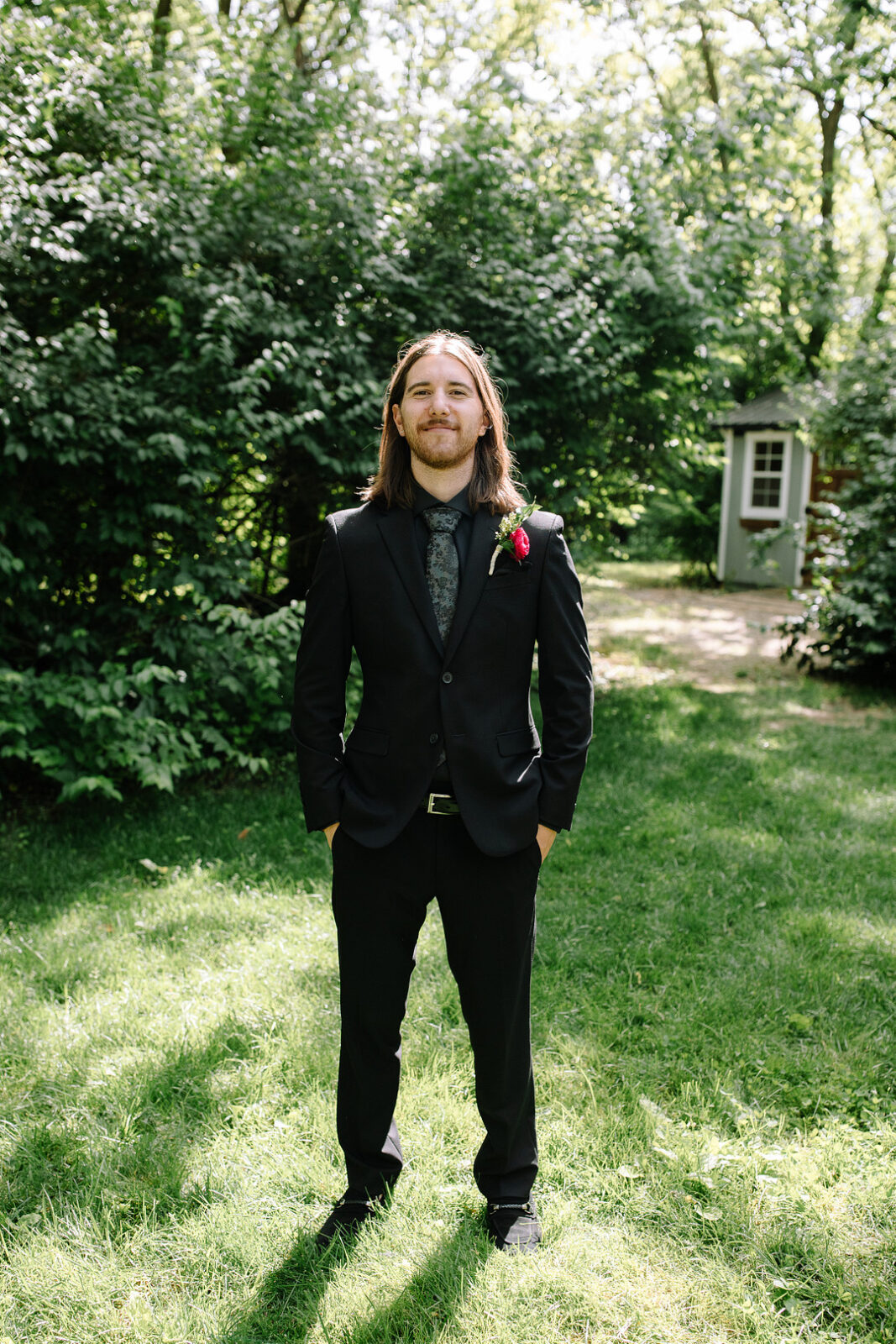 A man with long hair stands outdoors on grass at Stone Valley Meadows, wearing a black suit with a patterned tie and red boutonniere—perfectly dressed for a Spring Woodland Wedding. Green bushes and a small shed are in the background.
