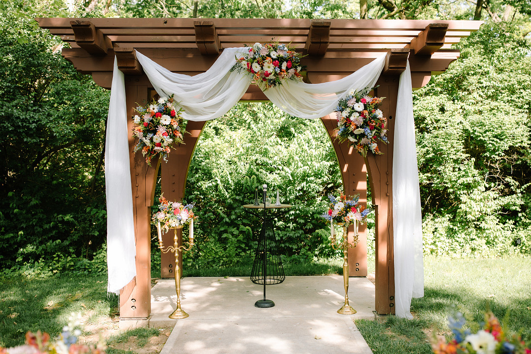 A wooden pergola decorated with white fabric and colorful floral arrangements stands on a concrete path surrounded by greenery at Stone Valley Meadows, perfect for a romantic Spring Woodland Wedding with a small round table underneath.