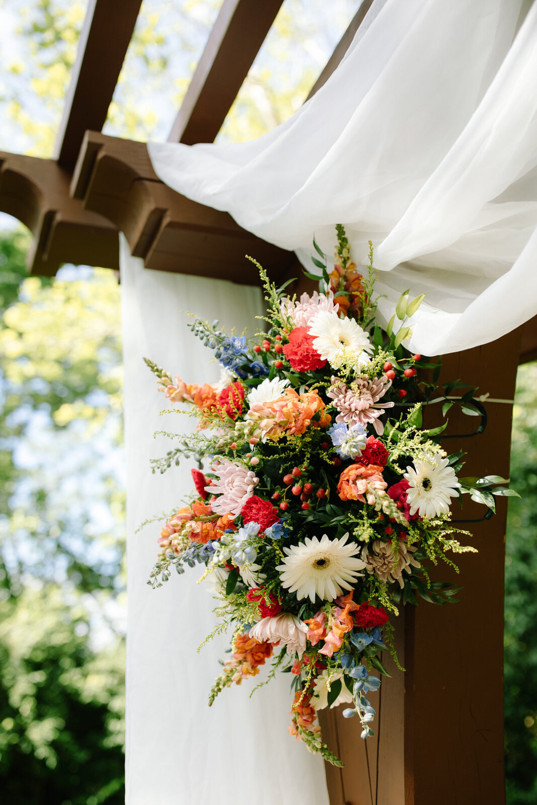A colorful floral arrangement featuring white, pink, orange, red, and blue flowers is attached to a wooden pergola draped with white fabric outdoors at Stone Valley Meadows for a stunning Spring Woodland Wedding.