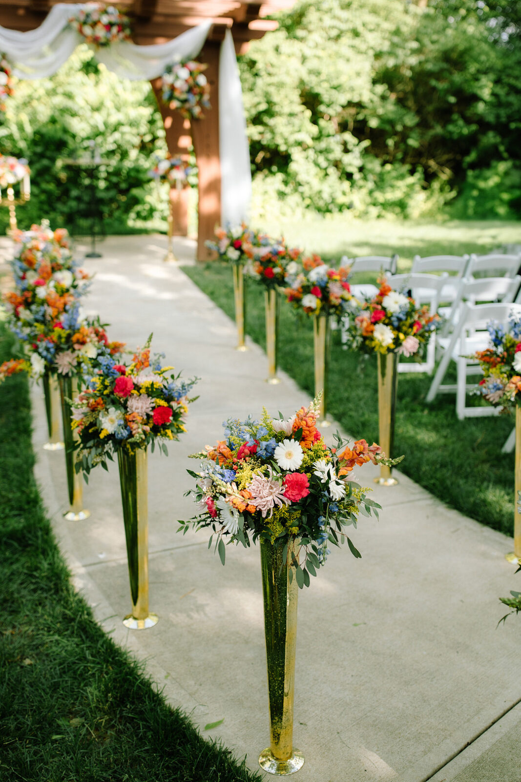 At Stone Valley Meadows, a Spring Woodland Wedding comes to life with an outdoor aisle lined by tall gold vases of vibrant flowers, leading to an altar draped in white and adorned with blooms. White chairs are perfectly arranged on either side.