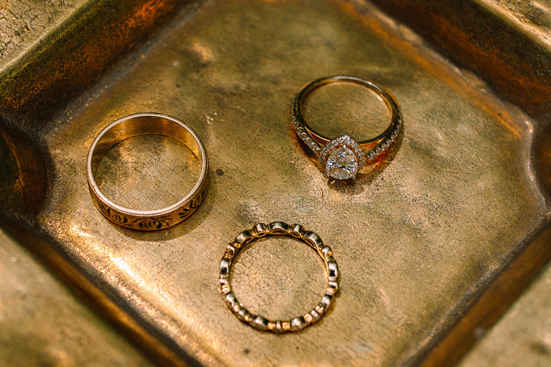 Three rings—a gold band, a diamond engagement ring, and a textured gold ring—are elegantly displayed on a brass tray, capturing the natural romance of a Spring Woodland Wedding at Stone Valley Meadows.