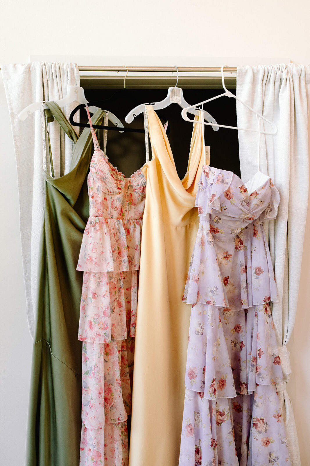 Four dresses on hangers hang from a curtain rod: an olive green dress, a yellow slip dress, and two tiered floral dresses in pink and lavender, perfect for a Spring Woodland Wedding at Stone Valley Meadows.