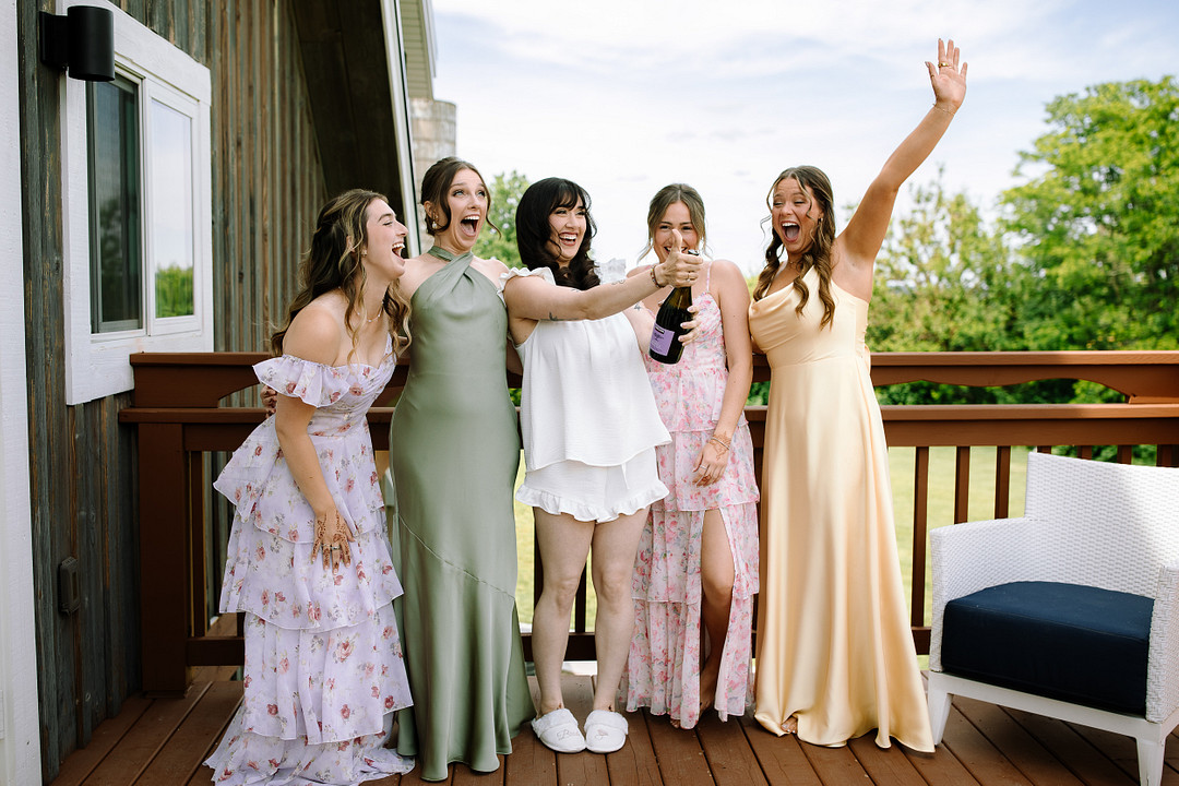 Five women stand on a deck at Stone Valley Meadows, smiling and holding a champagne bottle; four wear pastel dresses, one wears white loungewear and slippers—capturing the joy of a Spring Woodland Wedding celebration.