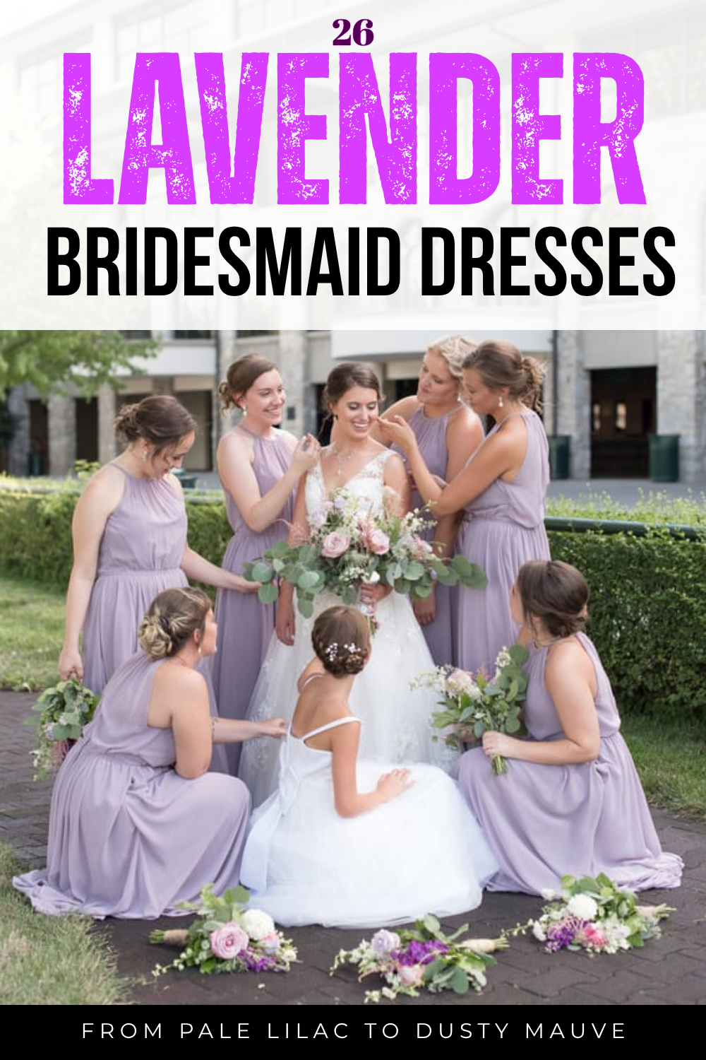 A bride in white is surrounded by eight bridesmaids wearing stylish lilac bridesmaid dresses, holding bouquets, outdoors near a building.
