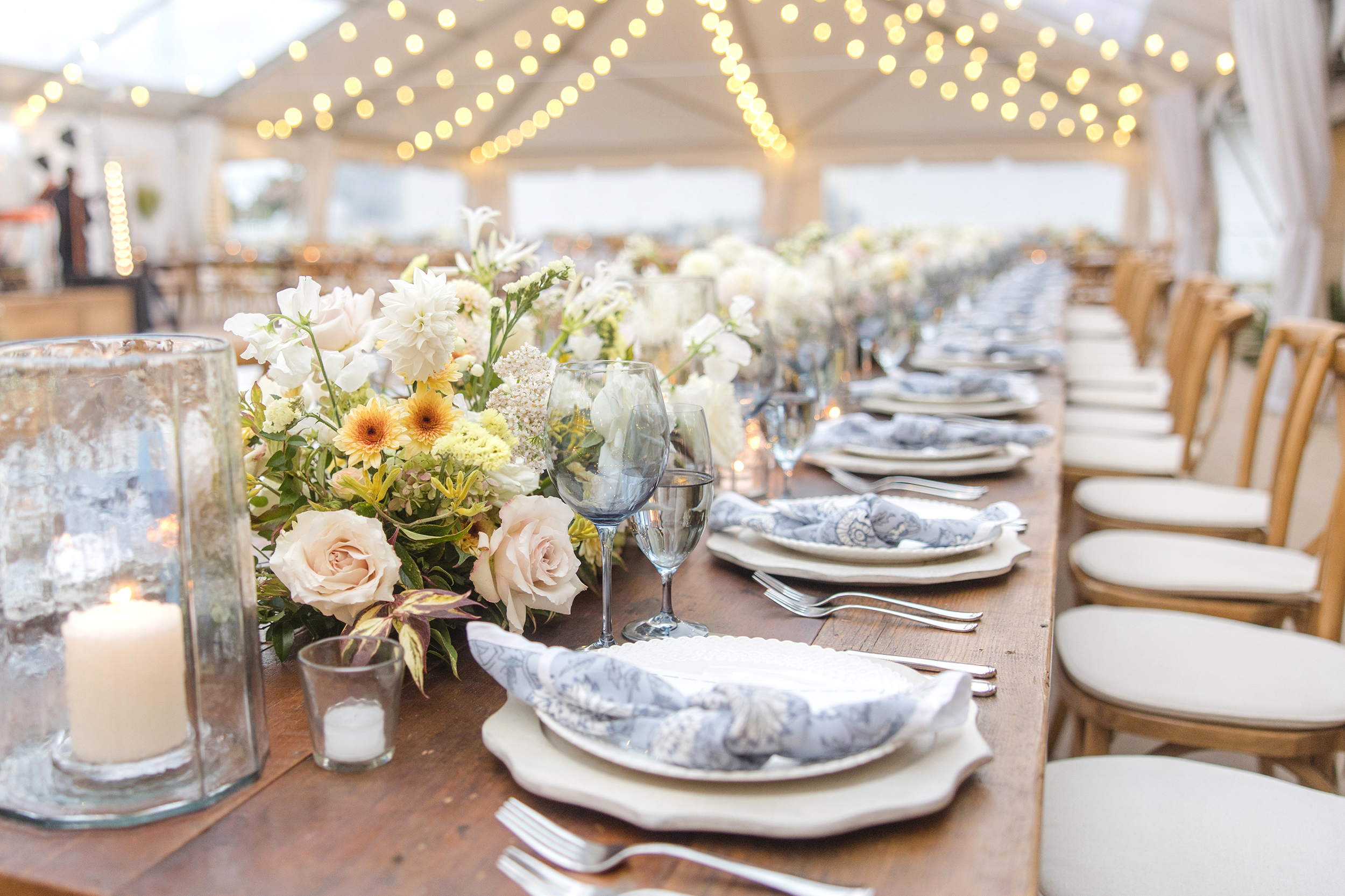 Long farm table under a clear-span tent with overhead string lights, white and yellow floral centerpieces, blue napkins, and candlelit place settings