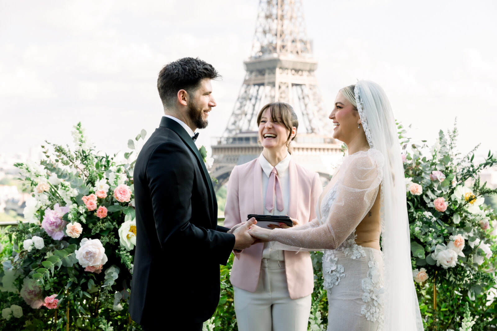 A couple stands holding hands during a wedding ceremony in front of the Eiffel Tower, with a smiling officiant and floral arrangements around them.