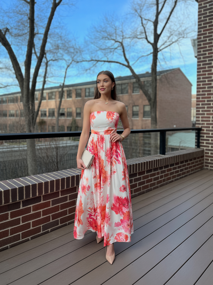 A woman in a strapless white spring wedding guest dress with red floral prints stands on a balcony, holding a clutch purse. Trees and a brick building are visible in the background.