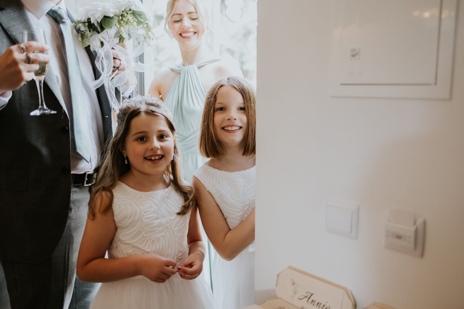 Two young girls in white dresses stand indoors, smiling at the camera, with adults and a flower bouquet partially visible behind them.