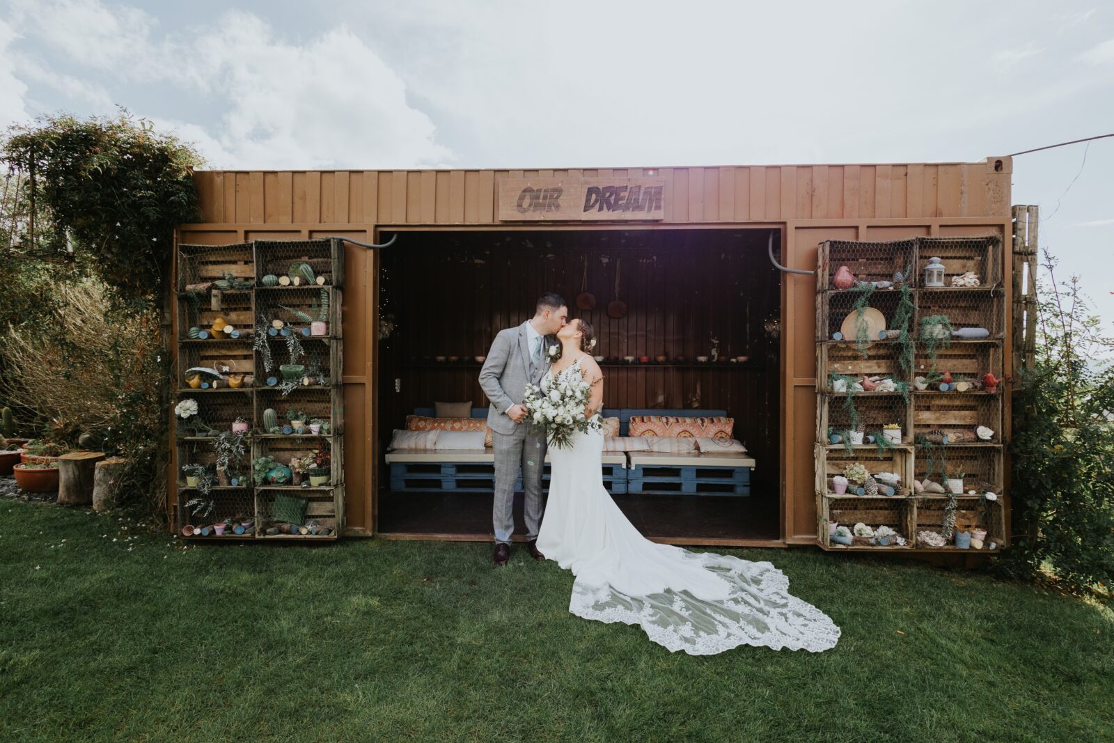 A bride and groom stand kissing in front of an open shipping container decorated with shelves of various objects and a sign reading "OUR DREAM.