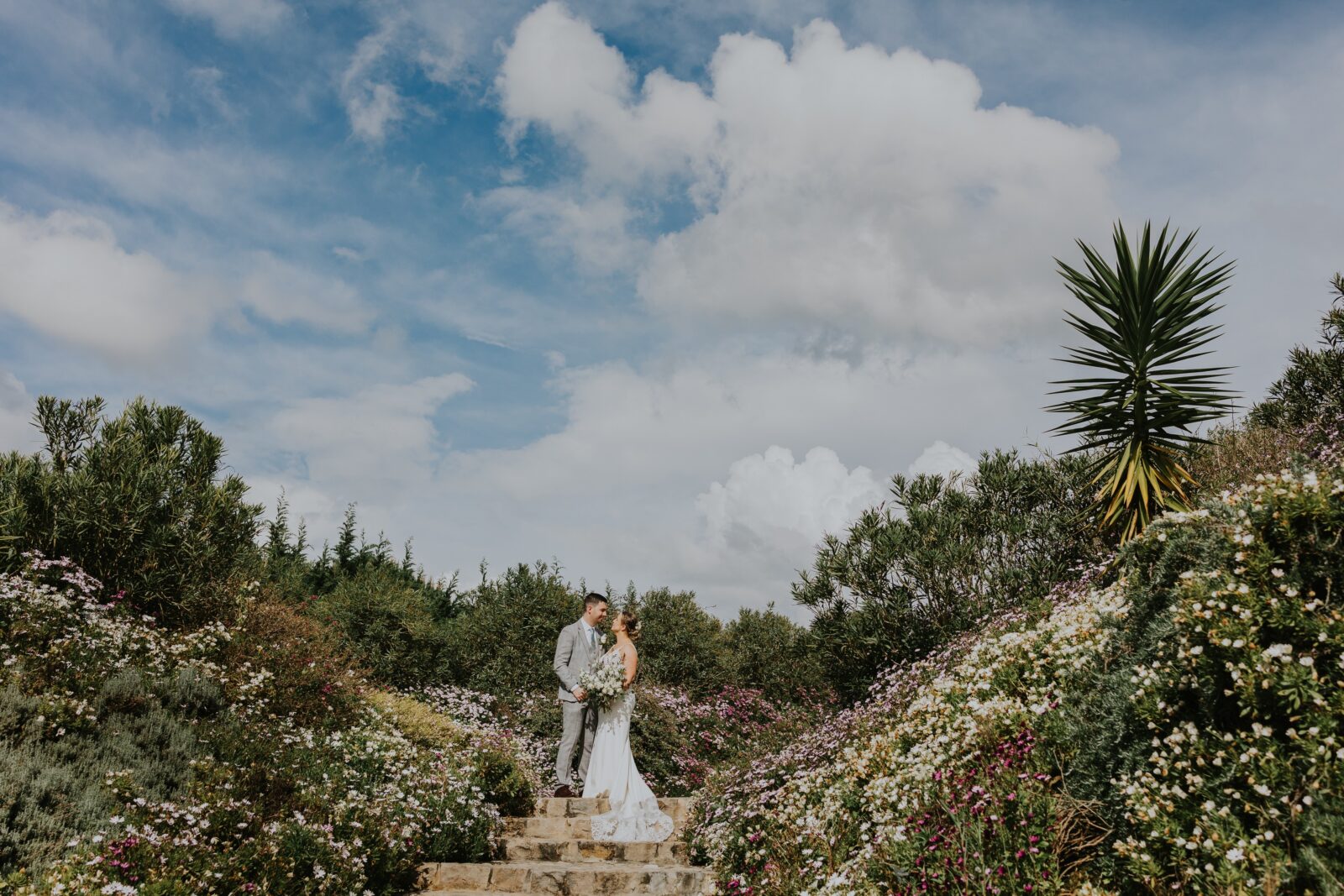 A bride and groom stand together on outdoor stone steps, surrounded by lush greenery and flowering plants under a partly cloudy sky.
