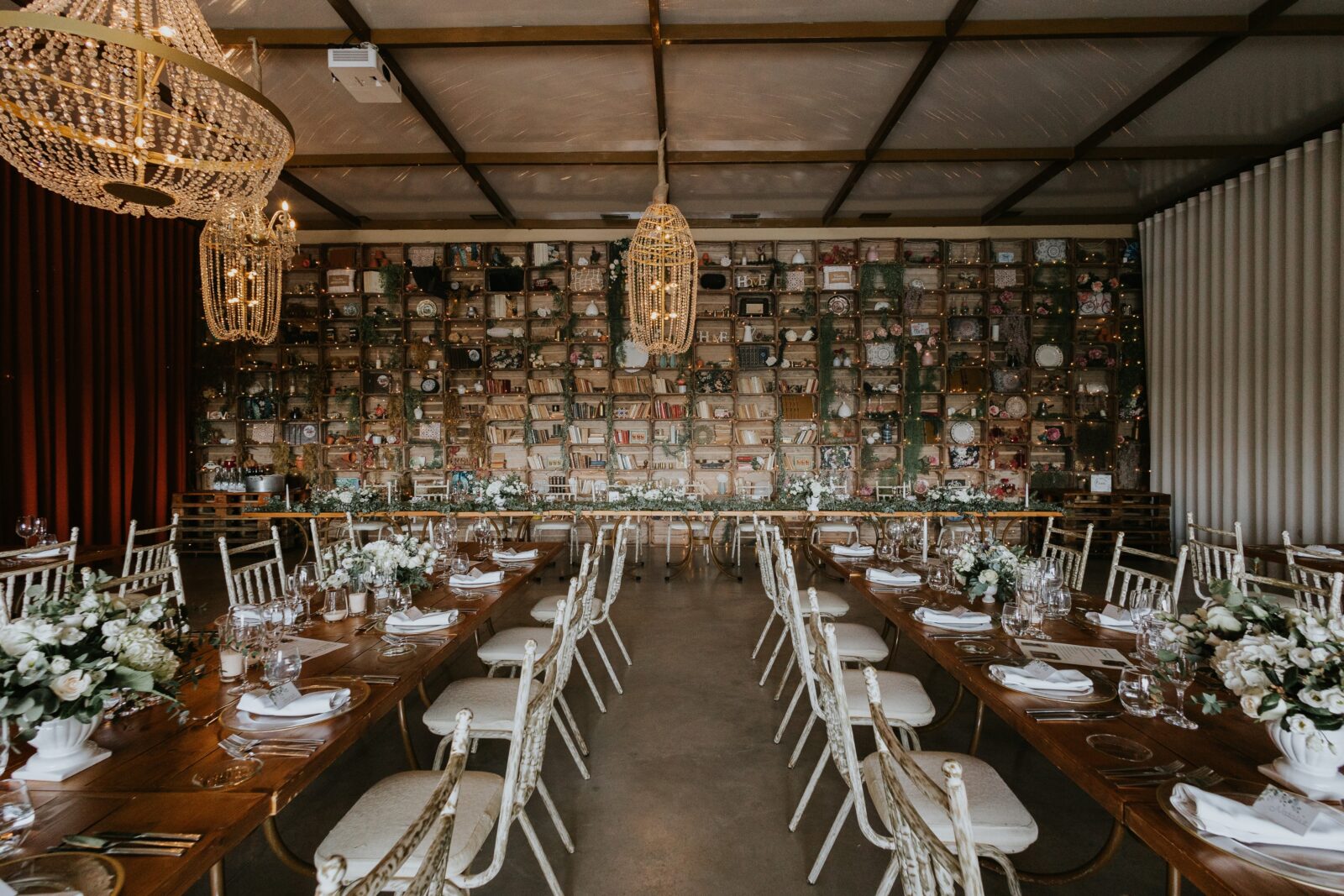 Elegant banquet hall with long wooden tables, white chairs, floral centerpieces, and two chandeliers, set against a wall decorated with vintage items and shelves.