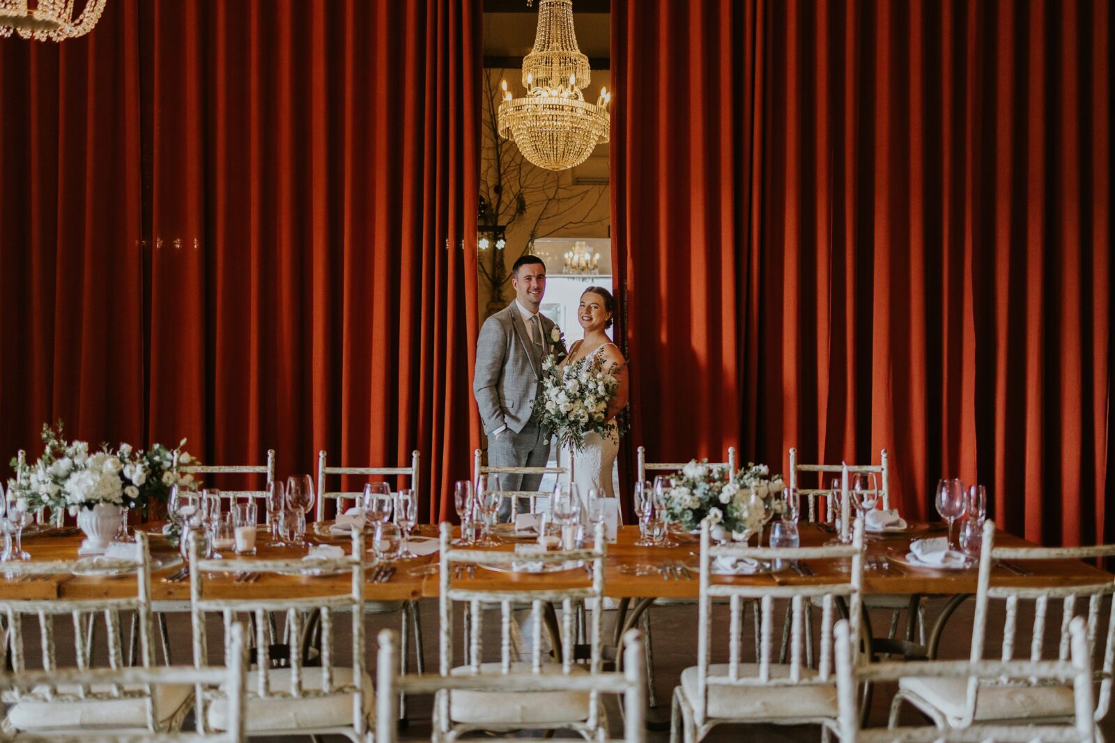Bride and groom stand in front of large red curtains behind a decorated banquet table set for a formal event.