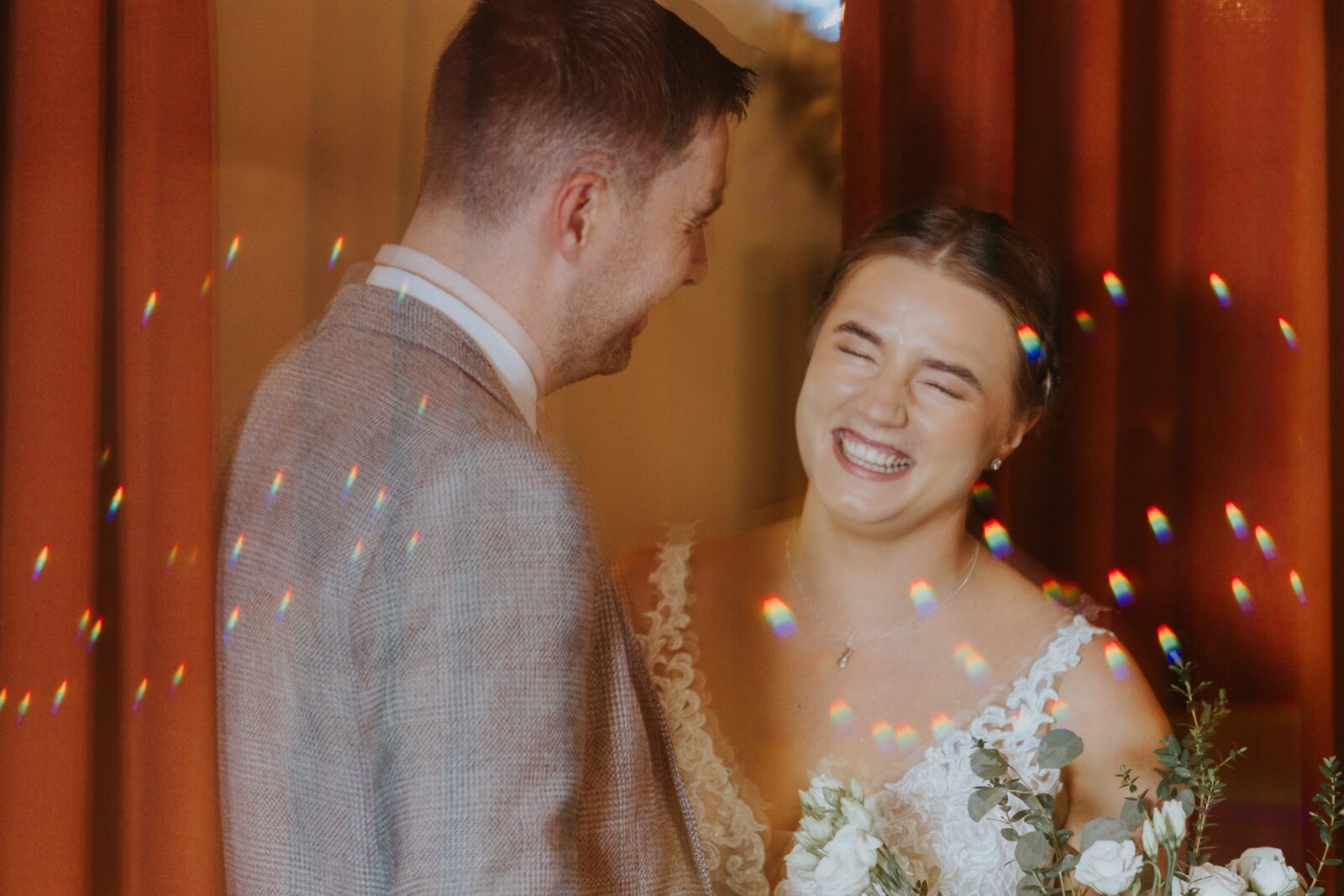 A bride and groom stand together indoors, smiling and laughing. The image features red curtains and scattered light reflections.