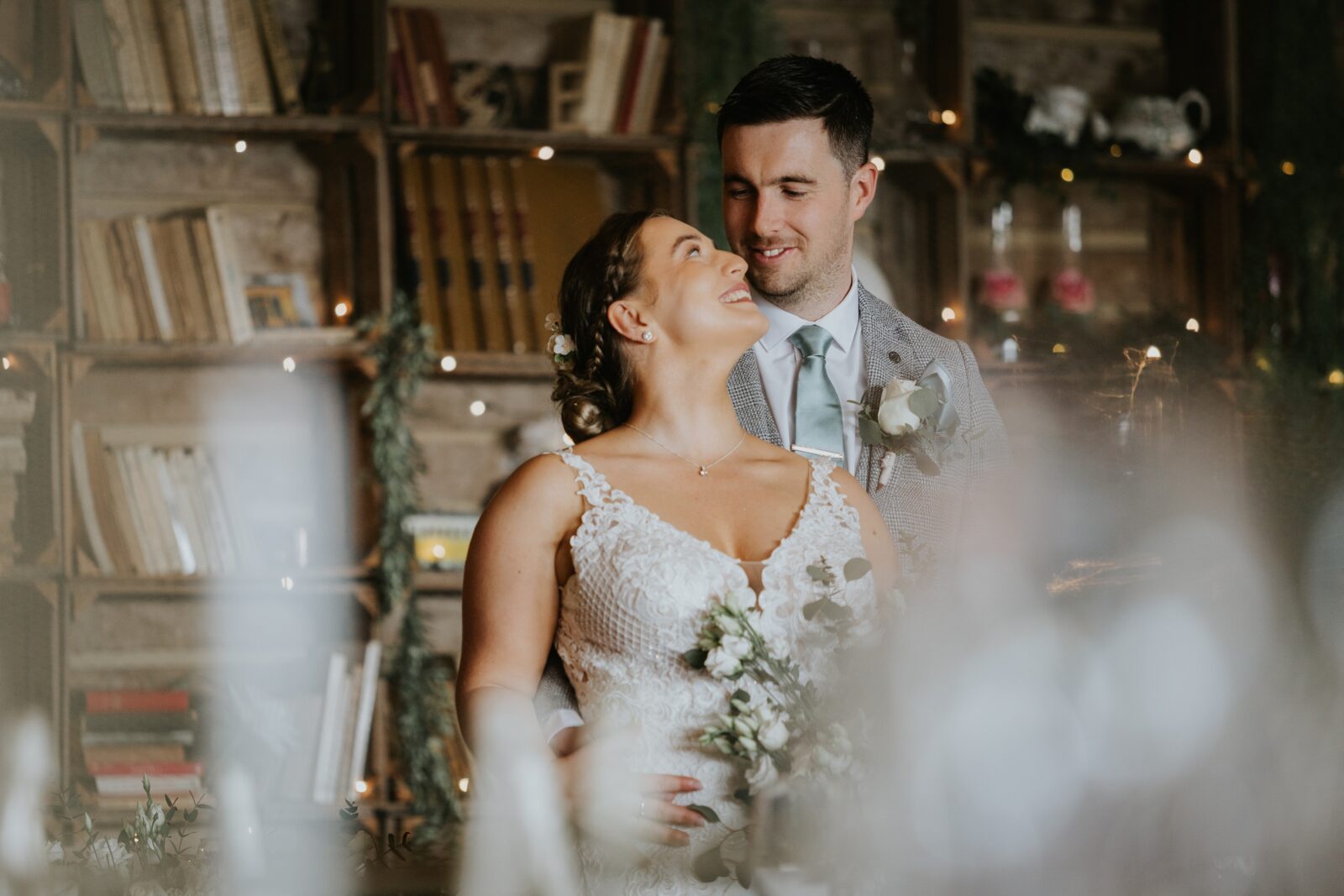 A bride and groom stand close together indoors, smiling and holding flowers, with bookshelves and soft decorative lighting in the background.