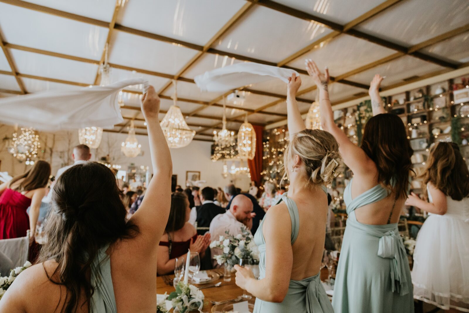 A group of women in light blue dresses wave white cloths in the air at a lively indoor wedding reception with chandeliers and decorated tables.