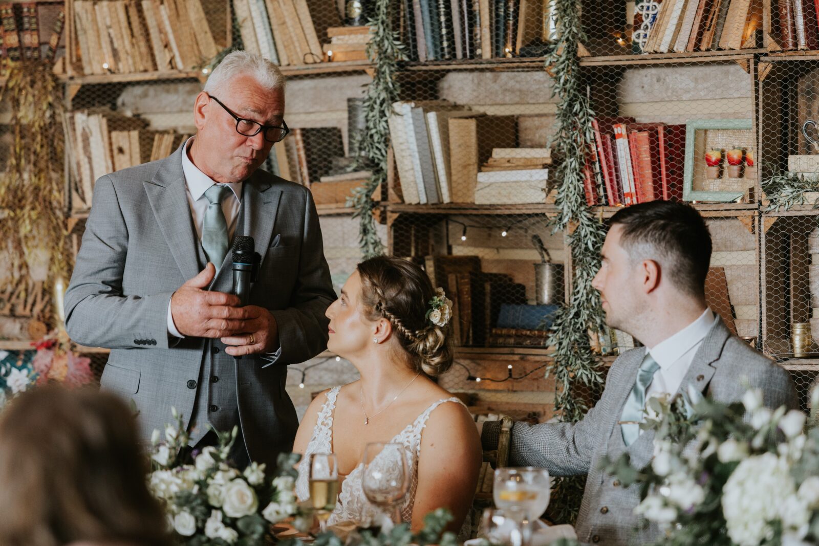 An older man in a grey suit speaks into a microphone to a seated bride and groom at a decorated table with bookshelves and flowers in the background.