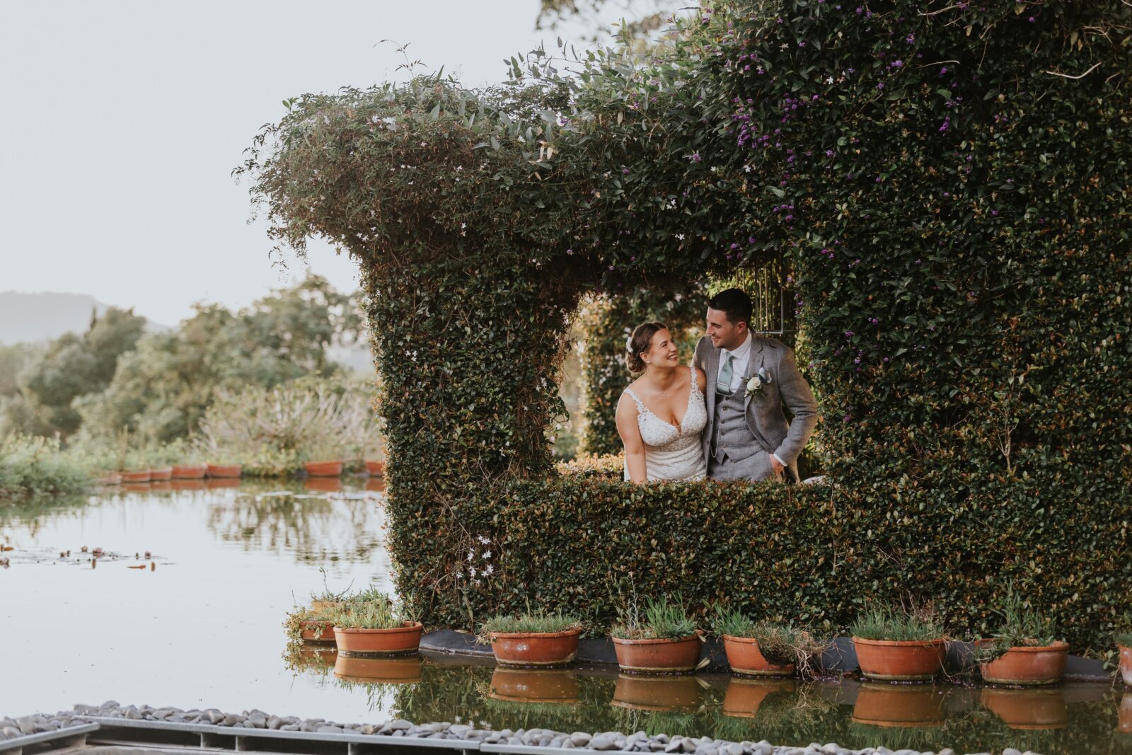 A bride and groom stand together inside a leafy, vine-covered structure beside a reflective pond, surrounded by potted plants.