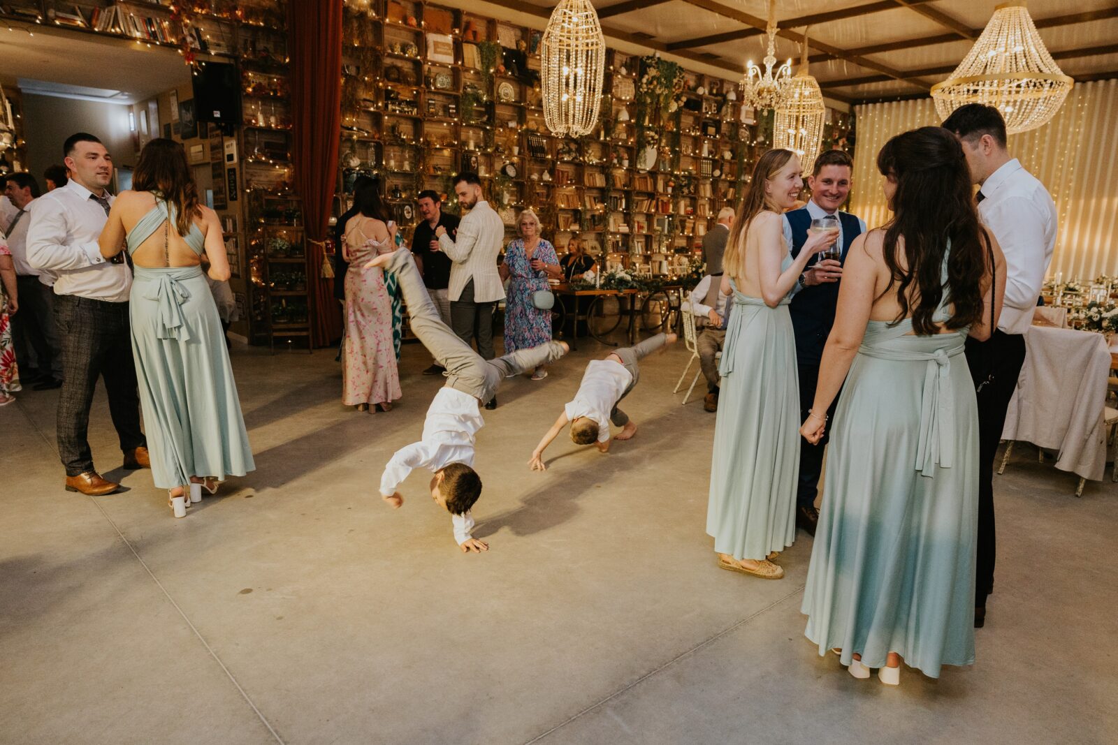 Two young boys do cartwheels on the dance floor at a formal indoor event while guests in formal attire stand and talk nearby.