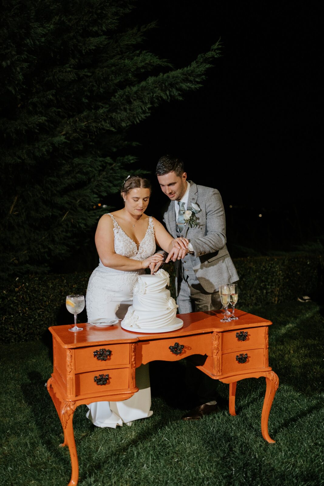 A bride and groom cut a white wedding cake together on an orange table outdoors at night.