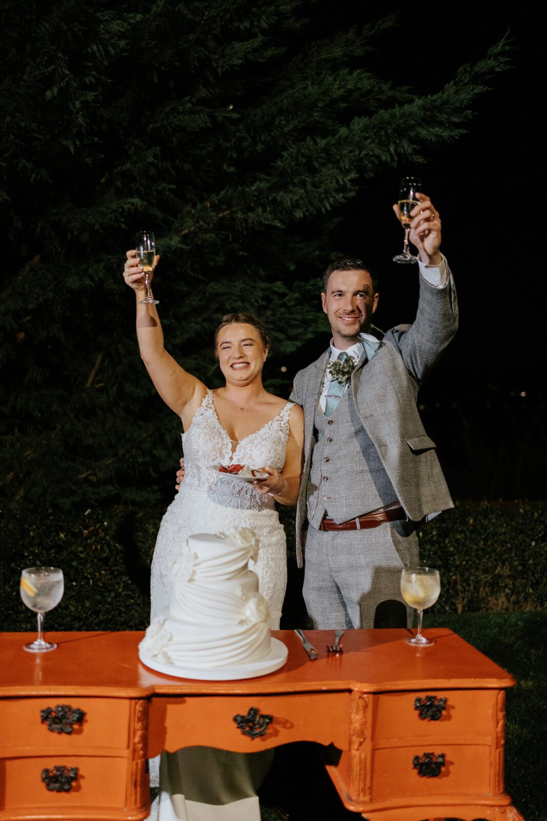 A bride and groom stand behind a cake on an orange table, holding up glasses in a toast at an outdoor nighttime wedding celebration.