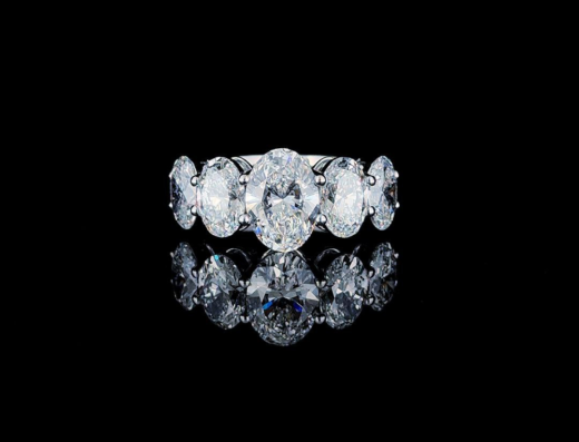 A silver ring with seven oval-cut diamonds is displayed against a black background, with its reflection visible below.