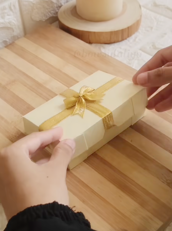 Hands holding a rectangular gift box wrapped in cream paper with a gold ribbon on a wooden surface, perfect for those seeking DIY Gift Box Ideas or inspiration for creative Gift Box Folding.