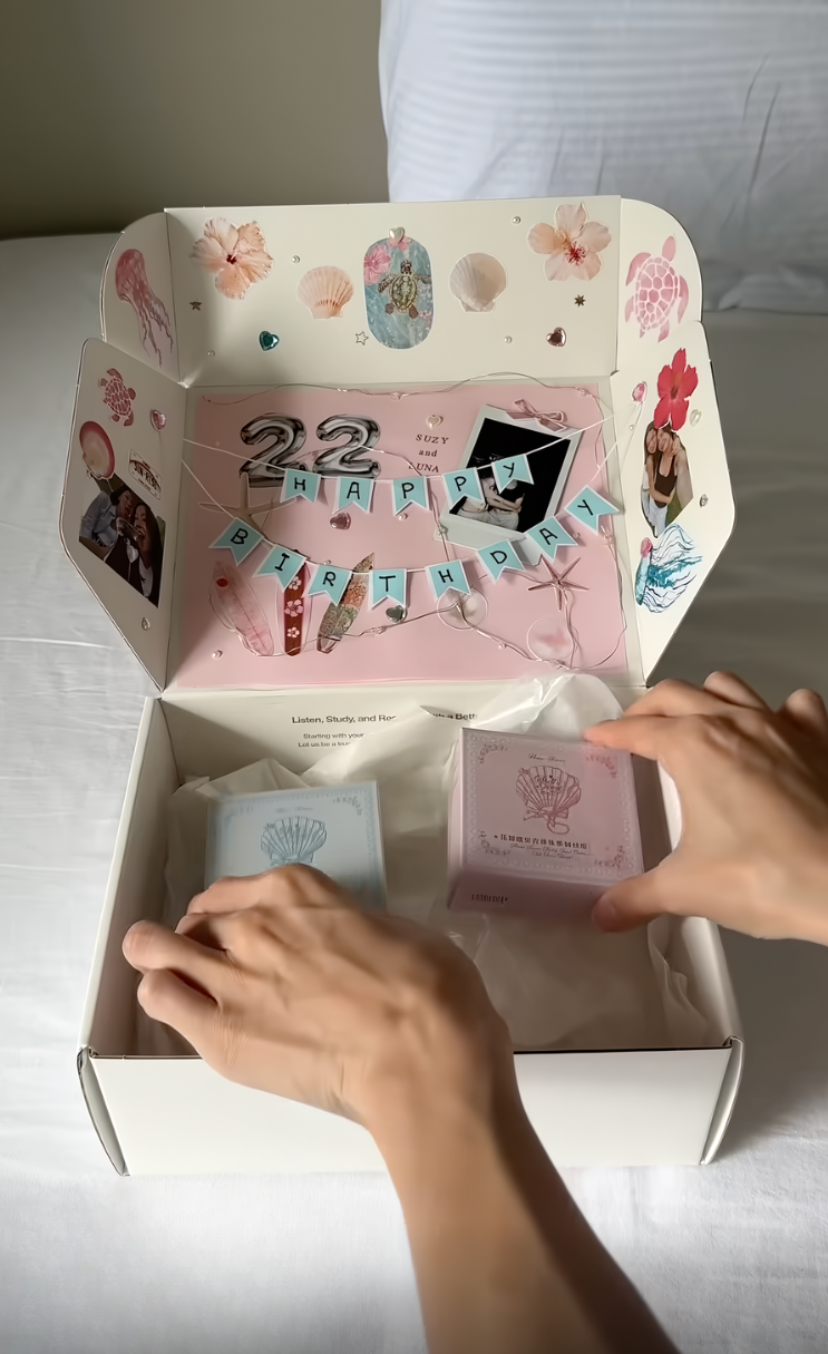 Open gift box on a bed with “Happy Birthday” decorations inside; two hands reach for pastel-colored items nestled in tissue paper—an inspiring example of DIY gift box ideas perfect for adding a personal touch to any special occasion.