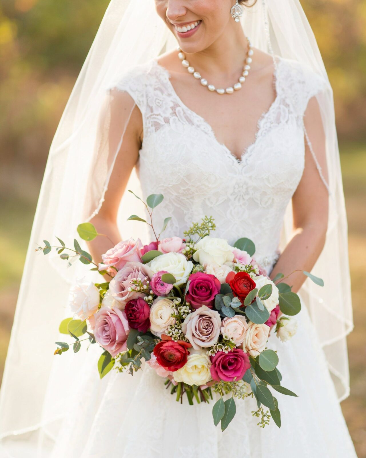 A bride in a white lace wedding dress and veil holds a bouquet of assorted pink, red, and white roses with greenery—perfect inspiration for Fall Wedding Flowers seen in Real Weddings on Pinterest.