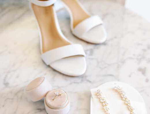 White heeled sandals, a velvet ring box with a diamond ring, and pearl drop earrings in packaging are arranged on a marble surface—perfect details for an Elegant Wedding at Cielo Farms.