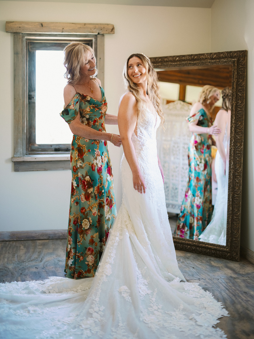 A woman in a floral dress helps a bride in a white gown adjust her dress in front of a large mirror, capturing an elegant wedding moment at Cielo Farms.
