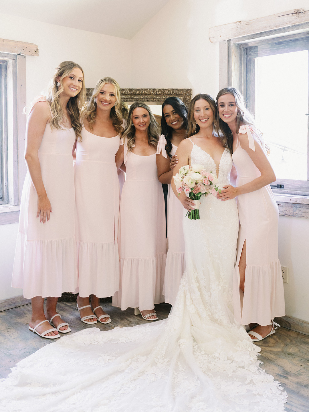 Five women stand indoors at Cielo Farms, four in matching light pink dresses and sandals, and one in a white wedding gown holding a bouquet—all smiling at the camera for this elegant wedding celebration.