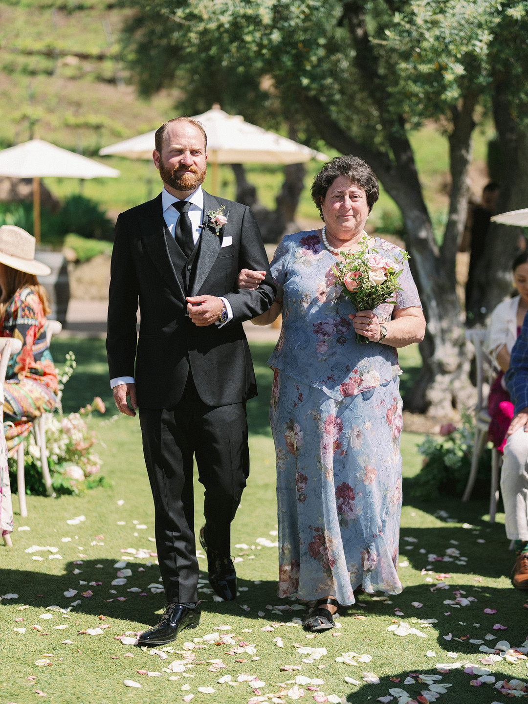 A man in a black suit walks arm-in-arm with a woman in a floral dress holding a bouquet at an elegant wedding outdoors on grass, with flower petals scattered and guests seated in the background.