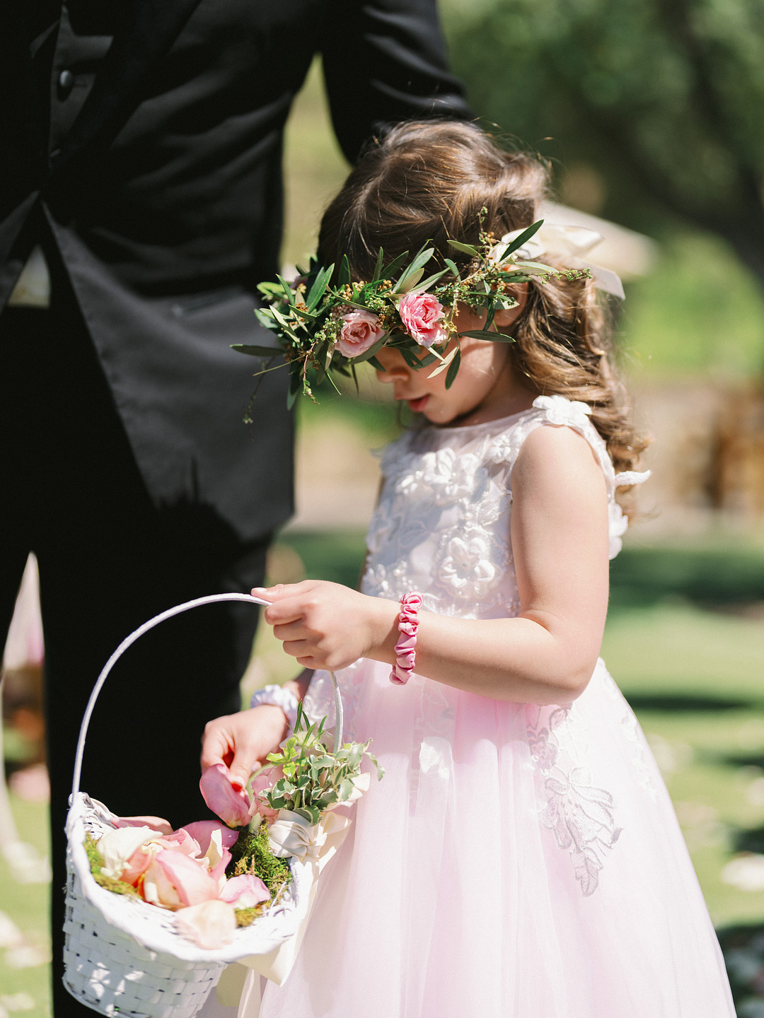 At an elegant wedding at Cielo Farms, a young girl in a white dress and floral crown holds a basket of flower petals outdoors, with an adult in a black suit standing beside her.
