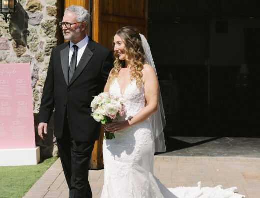 A bride in a white wedding dress holds a bouquet and walks beside an older man in a black suit outside Cielo Farms, an elegant wedding venue with stone buildings and large wooden doors.