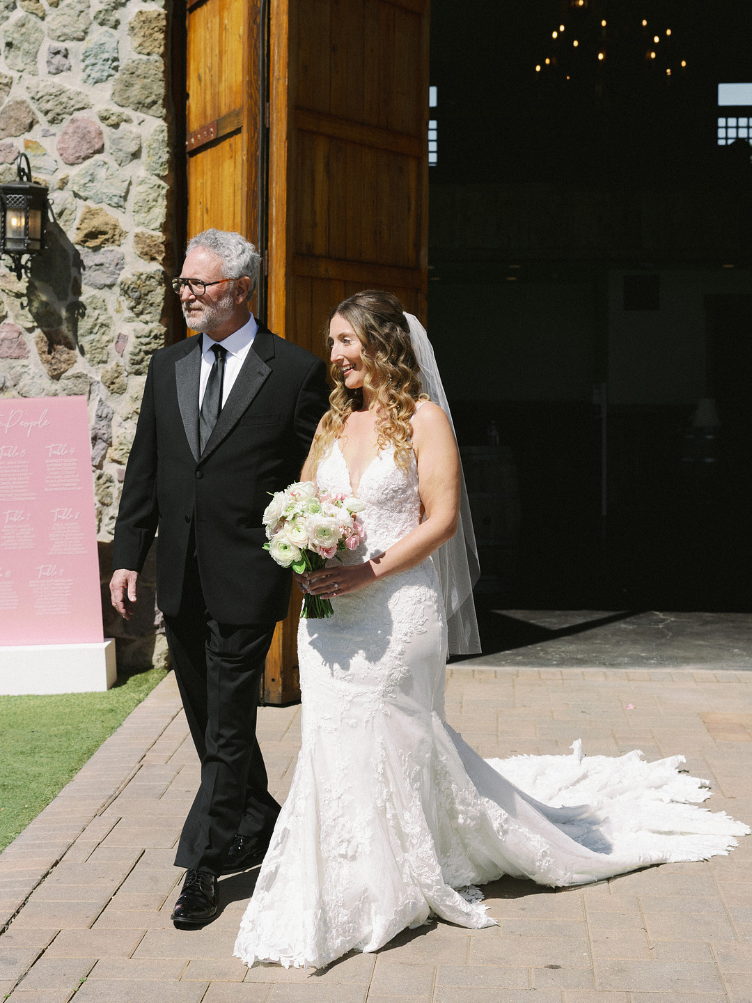 A bride in a white wedding dress holds a bouquet and walks beside an older man in a black suit outside Cielo Farms, an elegant wedding venue with stone buildings and large wooden doors.