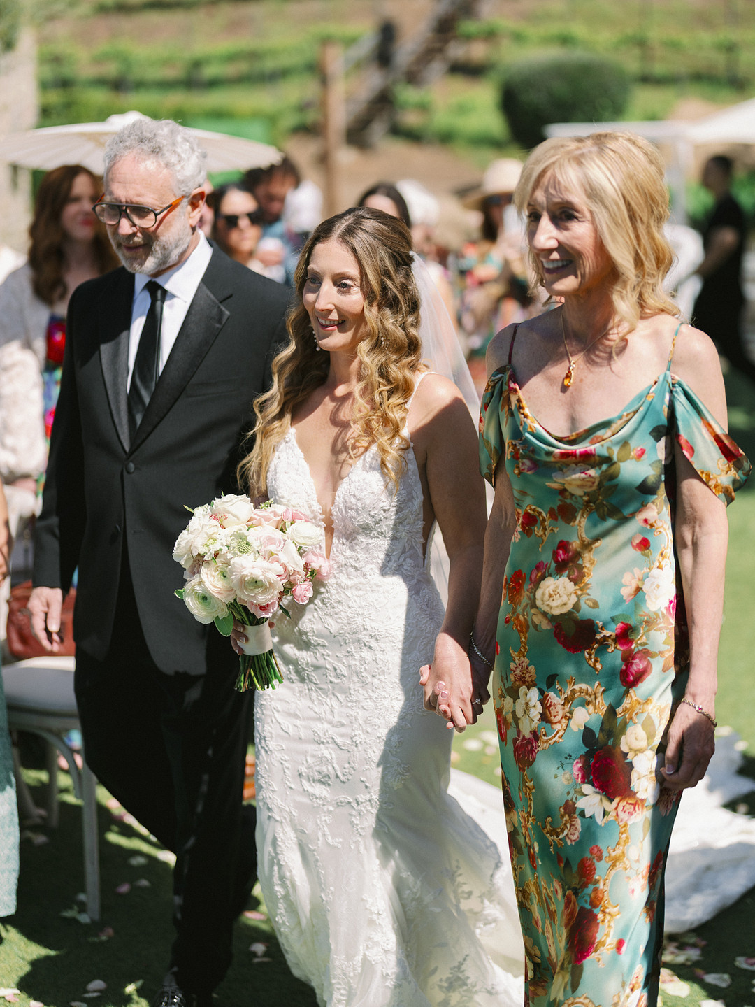A bride in a white lace gown holds a bouquet and walks outdoors at Cielo Farms, flanked by an older man in a suit and an older woman in a floral dress, capturing the charm of an elegant wedding.