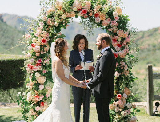 A bride and groom stand facing each other holding hands during an elegant wedding ceremony at Cielo Farms, with an officiant behind them and a large floral arch in the background.