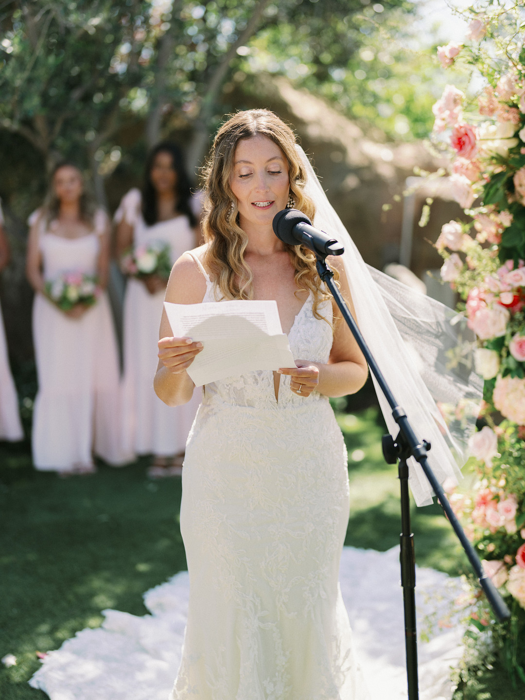 A bride in a white dress stands at a microphone outdoors at Cielo Farms, reading from a sheet of paper, with bridesmaids and floral arrangements adding to the elegant wedding atmosphere.