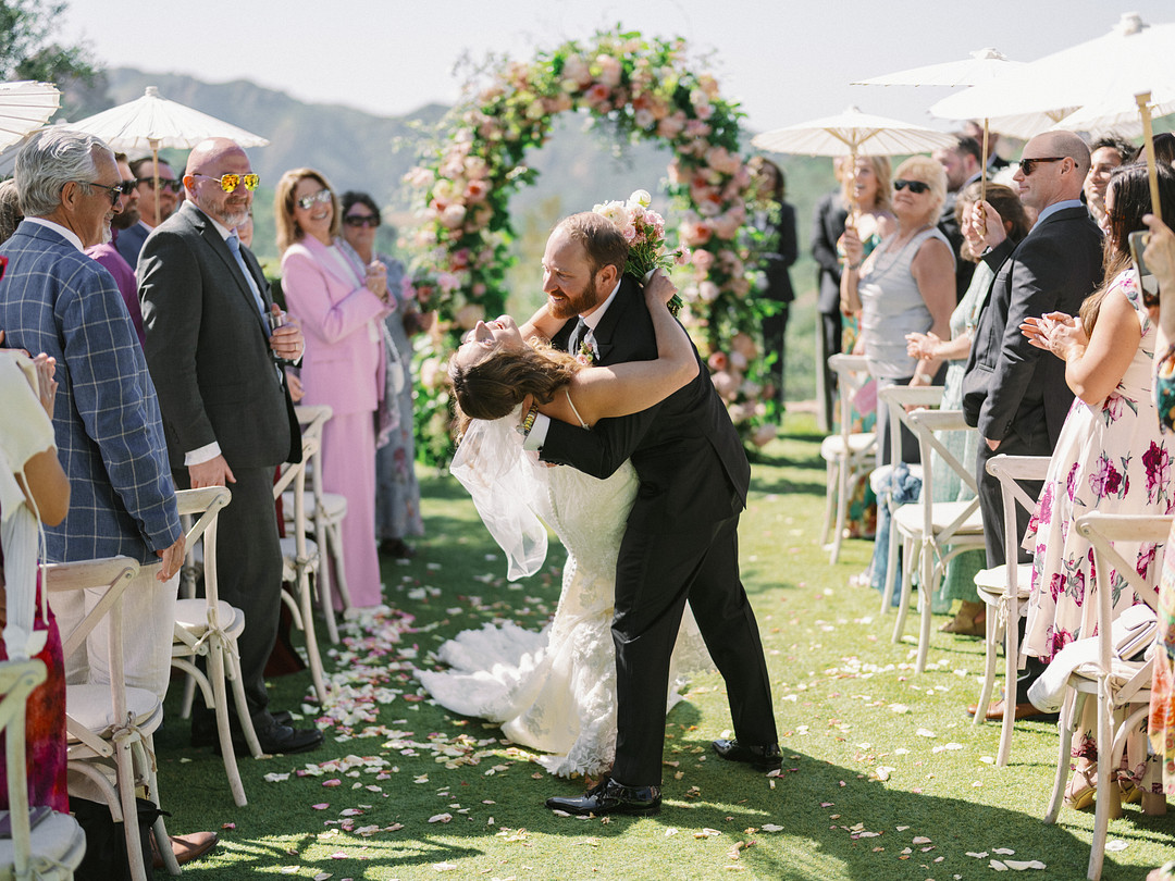 At an elegant spring wedding at Cielo Farms, a groom dips his bride for a kiss during the outdoor ceremony, surrounded by guests seated on either side of a flower-petal-lined aisle.