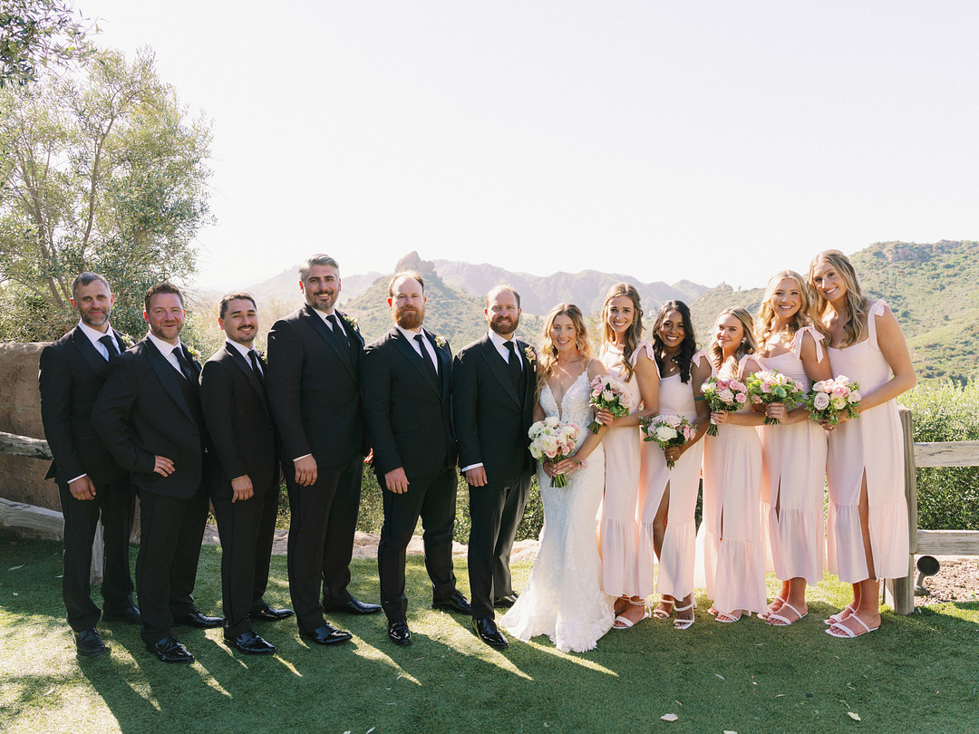 A wedding party stands outdoors at Cielo Farms in formal attire, with five men in dark suits and five women in light pink dresses posing on grass with mountains in the background, capturing an elegant wedding moment.